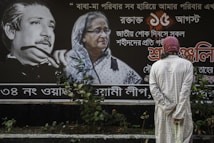 A person wearing a light-colored robe and a red and white headscarf stands in front of a large poster featuring two prominently displayed portraits accompanied by text in a script that could be Bengali. The portrait on the left shows an older man with distinctive facial features, while the portrait on the right shows an older woman wearing glasses and a white headscarf. The background of the poster is predominantly dark, with white text and accents in red.