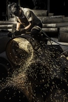 A worker focuses intently while using a grinding tool, with bright sparks flying from a large metal pipe. The setting appears to be an industrial area with various metal pipes stacked in the background.