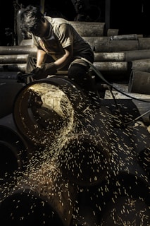 A worker focuses intently while using a grinding tool, with bright sparks flying from a large metal pipe. The setting appears to be an industrial area with various metal pipes stacked in the background.