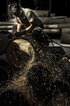 A worker focuses intently while using a grinding tool, with bright sparks flying from a large metal pipe. The setting appears to be an industrial area with various metal pipes stacked in the background.