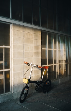 A compact folding bicycle with a black frame, orange handlebars, and a red saddle leans against a concrete block wall, which is partially illuminated by soft sunlight. Large windows with grid patterns line the wall, casting subtle reflections and shadows onto the surface.