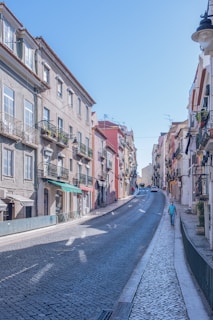 A narrow cobblestone street lined with colorful buildings with balconies and plants. The street ascends a hill, and there is a single person walking uphill. Cars are parked along the road, and the sun casts shadows across the pathway.