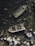 Two abandoned boats lie partially submerged in shallow water surrounded by rocks. The boats appear weathered and filled with debris, with one being more deteriorated than the other. The surrounding area is rocky, with sharp and jagged stones visible around the vessels.