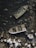 Two abandoned boats lie partially submerged in shallow water surrounded by rocks. The boats appear weathered and filled with debris, with one being more deteriorated than the other. The surrounding area is rocky, with sharp and jagged stones visible around the vessels.