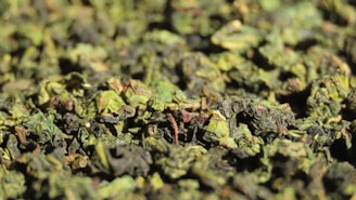 A close-up view of a pile of dried, rolled green tea leaves with a mixture of brown and green hues. The texture appears rough and organic, with various sizes of leaves clustered together.