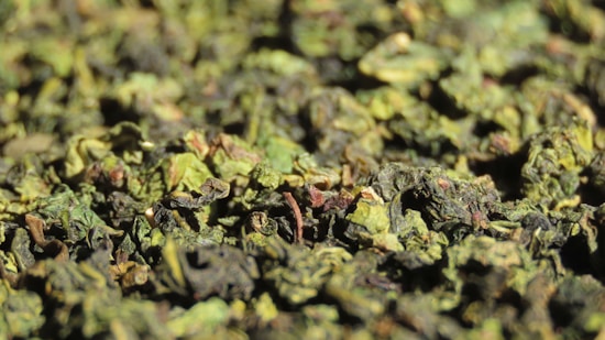 A close-up view of a pile of dried, rolled green tea leaves with a mixture of brown and green hues. The texture appears rough and organic, with various sizes of leaves clustered together.