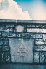 A stone wall with a large plaque featuring the outline of a tree and the words 'Hamilton County Park District'. The sky above is partly cloudy with a bright and clear blue section.