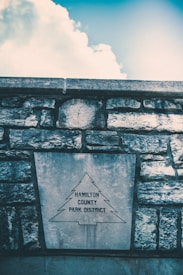 A stone wall with a large plaque featuring the outline of a tree and the words 'Hamilton County Park District'. The sky above is partly cloudy with a bright and clear blue section.