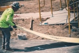 A construction worker wearing a safety helmet and a bright green long-sleeve shirt is handling a long piece of yellow board on a site. The background shows dirt, scaffolding, and construction materials, indicating an ongoing building process.