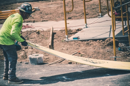 A construction worker wearing a safety helmet and a bright green long-sleeve shirt is handling a long piece of yellow board on a site. The background shows dirt, scaffolding, and construction materials, indicating an ongoing building process.