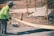 A construction worker wearing a safety helmet and a bright green long-sleeve shirt is handling a long piece of yellow board on a site. The background shows dirt, scaffolding, and construction materials, indicating an ongoing building process.