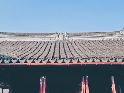 A traditional roof with curved ceramic tiles is set against a clear blue sky. The roof features ornate sculptural details depicting figures along the ridge. Red wooden beams support the structure, suggesting traditional architecture.