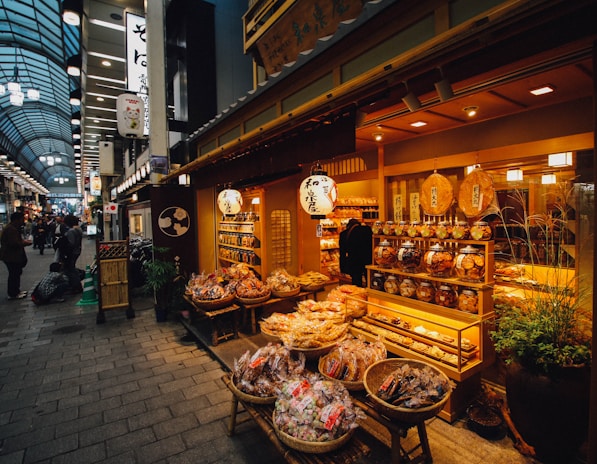 A bustling indoor market scene featuring a traditional Japanese store. The store displays an array of packaged snacks and goods in wicker baskets and wooden shelves. The warm lighting highlights the rich colors of the products. Shoppers pass by and explore the market, showcasing a vibrant and lively atmosphere.