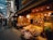 A bustling indoor market scene featuring a traditional Japanese store. The store displays an array of packaged snacks and goods in wicker baskets and wooden shelves. The warm lighting highlights the rich colors of the products. Shoppers pass by and explore the market, showcasing a vibrant and lively atmosphere.