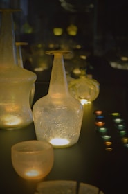 A collection of translucent glass bottles and cups illuminated by soft, warm light. The glassware displays a textured surface, highlighted against a dark background. Small, colorful round objects are arranged neatly to the side, adding contrast.