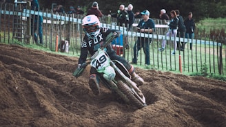 A motorcyclist in full gear navigates a dirt track, kicking up soil as they lean into a turn. Spectators stand behind a wooden fence, observing the race. The rider wears a helmet, goggles, and a motocross jersey with the number '49' visible on the bike.