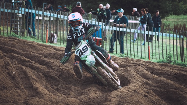 A motorcyclist in full gear navigates a dirt track, kicking up soil as they lean into a turn. Spectators stand behind a wooden fence, observing the race. The rider wears a helmet, goggles, and a motocross jersey with the number '49' visible on the bike.