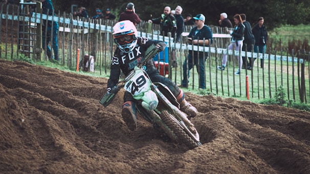 A motorcyclist in full gear navigates a dirt track, kicking up soil as they lean into a turn. Spectators stand behind a wooden fence, observing the race. The rider wears a helmet, goggles, and a motocross jersey with the number '49' visible on the bike.