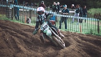 A motorcyclist in full gear navigates a dirt track, kicking up soil as they lean into a turn. Spectators stand behind a wooden fence, observing the race. The rider wears a helmet, goggles, and a motocross jersey with the number '49' visible on the bike.