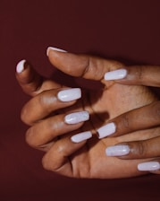 A hand with well-manicured nails painted in a pale pink color, with some nails featuring a glittery finish. The background is a solid deep brown, emphasizing the hand and nail design.