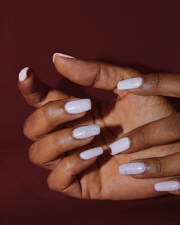A hand with well-manicured nails painted in a pale pink color, with some nails featuring a glittery finish. The background is a solid deep brown, emphasizing the hand and nail design.