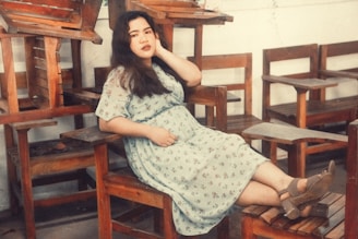 A person in a floral dress lounges comfortably on a wooden chair, surrounded by a stack of other wooden chairs and desks. The setting suggests a relaxed, perhaps contemplative, atmosphere in an indoor space with a warm color palette.