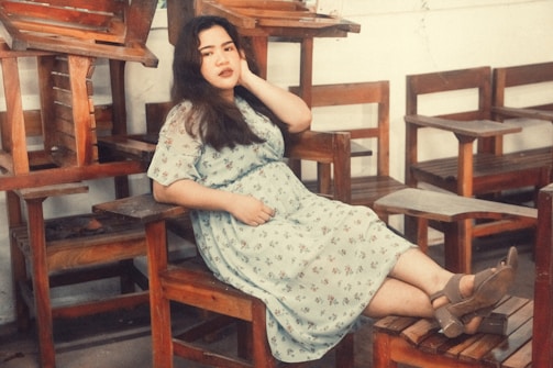 A person in a floral dress lounges comfortably on a wooden chair, surrounded by a stack of other wooden chairs and desks. The setting suggests a relaxed, perhaps contemplative, atmosphere in an indoor space with a warm color palette.