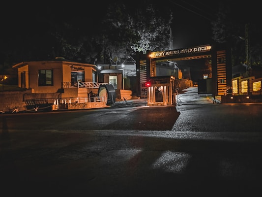 A nighttime scene depicting the entrance to a facility labeled as the Army School of Logistics. The area is dimly lit, with streetlights casting shadows. The entrance is marked by a decorative gate and guard hut. The atmosphere appears quiet and secure.