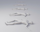 Several identical human figures are positioned in a line, lying down on a smooth, gradient surface that transitions from light gray to white. They appear to be sculptural or mannequin-like, with each figure slightly smaller and more distant than the previous one.