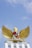 A golden eagle with its wings spread wide stands atop a white structure. Its chest is adorned with a shield containing various symbols. Below the eagle is an inscribed banner. The sky is clear and blue, providing a serene backdrop.