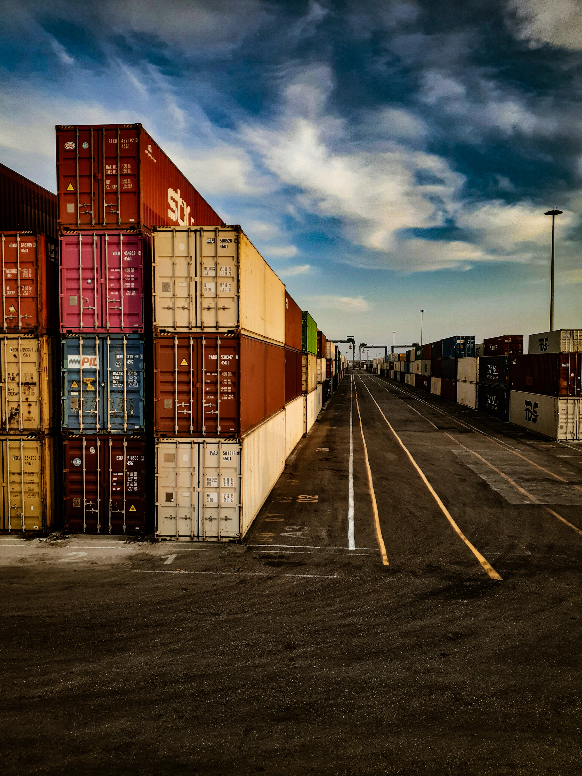 A container yard lane between walls of stacked shipping containers under a dramatic sky