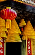 A series of golden-hued spiral incense coils hang in front of a traditional structure with intricate detailing. A vibrant red lantern with gold accents complements the scene. The backdrop includes Chinese characters on a vertical red banner.