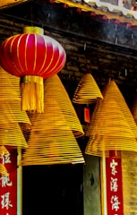 A series of golden-hued spiral incense coils hang in front of a traditional structure with intricate detailing. A vibrant red lantern with gold accents complements the scene. The backdrop includes Chinese characters on a vertical red banner.