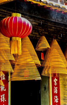 A series of golden-hued spiral incense coils hang in front of a traditional structure with intricate detailing. A vibrant red lantern with gold accents complements the scene. The backdrop includes Chinese characters on a vertical red banner.