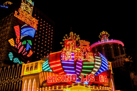 Bright neon lights and colorful signage adorn the exterior of Casino Lisboa, creating a vibrant and lively atmosphere. The façade features bold, multicolored designs with a mix of English and Chinese characters prominently displayed. The lights form intricate patterns and shapes, contributing to the energetic ambiance of the scene.