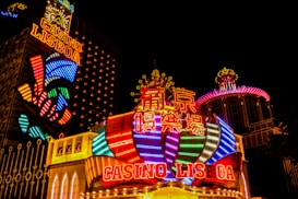 Bright neon lights and colorful signage adorn the exterior of Casino Lisboa, creating a vibrant and lively atmosphere. The fa&ccedil;ade features bold, multicolored designs with a mix of English and Chinese characters prominently displayed. The lights form intricate patterns and shapes, contributing to the energetic ambiance of the scene.