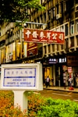 A street scene in a city featuring a prominent street sign labeled 'Avenida do Infante D. Henrique' with Chinese characters above. The sign is surrounded by greenery in an urban setting with a background of multi-story building façades displaying multiple store signs, including one in red featuring Chinese characters, indicating a commercial area. Pedestrians can be seen walking along the sidewalk, contributing to the bustling atmosphere.