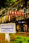 A street scene in a city featuring a prominent street sign labeled 'Avenida do Infante D. Henrique' with Chinese characters above. The sign is surrounded by greenery in an urban setting with a background of multi-story building façades displaying multiple store signs, including one in red featuring Chinese characters, indicating a commercial area. Pedestrians can be seen walking along the sidewalk, contributing to the bustling atmosphere.