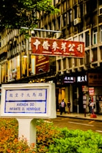 A street scene in a city featuring a prominent street sign labeled 'Avenida do Infante D. Henrique' with Chinese characters above. The sign is surrounded by greenery in an urban setting with a background of multi-story building façades displaying multiple store signs, including one in red featuring Chinese characters, indicating a commercial area. Pedestrians can be seen walking along the sidewalk, contributing to the bustling atmosphere.