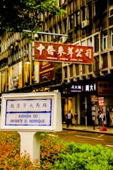 A street scene in a city featuring a prominent street sign labeled 'Avenida do Infante D. Henrique' with Chinese characters above. The sign is surrounded by greenery in an urban setting with a background of multi-story building façades displaying multiple store signs, including one in red featuring Chinese characters, indicating a commercial area. Pedestrians can be seen walking along the sidewalk, contributing to the bustling atmosphere.