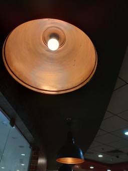 A warm, glowing ceiling lamp with an industrial design is surrounded by a dark ceiling, paired with a similar lamp visible in the background. The setting appears to be a modern indoor space, possibly a cafe or restaurant. The ceiling features a grid pattern, with recessed lighting providing additional illumination.