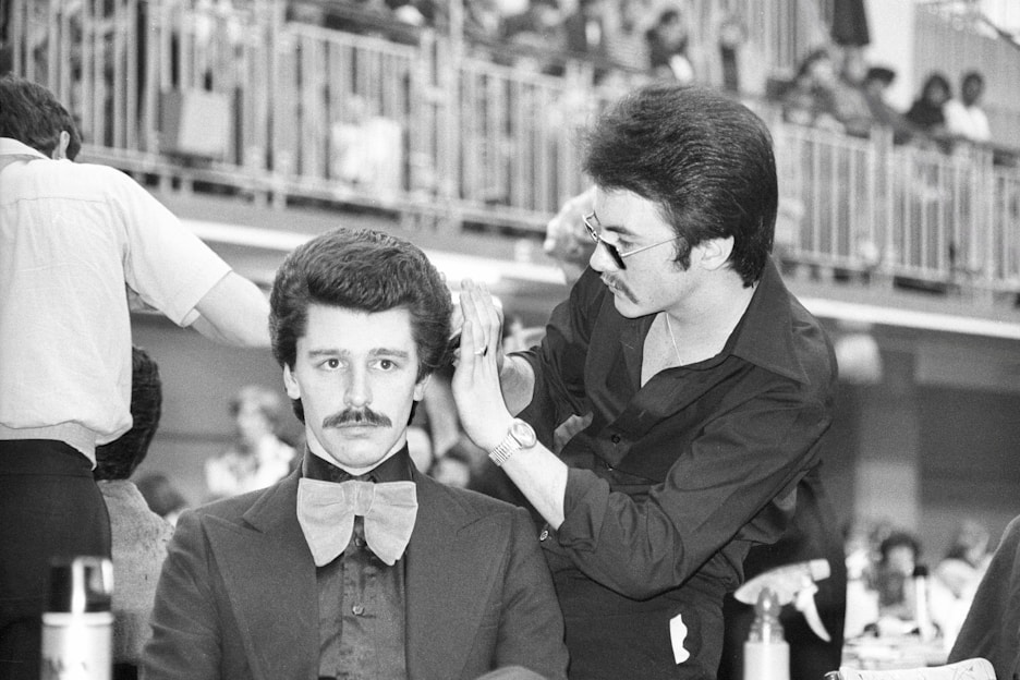 A man wearing glasses and a black shirt styles another man's hair, who is wearing a suit and a large bow tie, possibly in a public event setting. The background shows an audience seated behind railings, suggesting an event or competition.