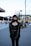 A person with a gothic style stands outdoors in an urban setting. They are wearing a black outfit, including a lace top, jacket, and pants, accessorized with a necklace featuring a cross. The background shows a city scene with people walking, modern buildings, and construction cranes.