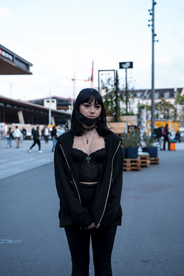 A striking model dressed in dark gothic attire standing against a shadowy urban backdrop.