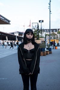 A person with a gothic style stands outdoors in an urban setting. They are wearing a black outfit, including a lace top, jacket, and pants, accessorized with a necklace featuring a cross. The background shows a city scene with people walking, modern buildings, and construction cranes.