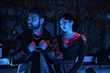 A man and a woman are standing together in a dimly lit setting with intense blue and red lighting. The man wears a dark leather jacket and holds onto a chain around his neck, while the woman, adorned in a dark dress with a necklace, gazes downward. Both individuals have a contemplative and serious expression.