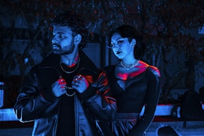 A man and a woman are standing together in a dimly lit setting with intense blue and red lighting. The man wears a dark leather jacket and holds onto a chain around his neck, while the woman, adorned in a dark dress with a necklace, gazes downward. Both individuals have a contemplative and serious expression.