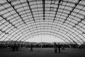 A large, open pavilion with an arched, transparent metal and glass roof structure. Several people, both standing and seated, are scattered throughout, with some in conversation and others facing the panoramic view of a cityscape and distant hills visible through the structure.