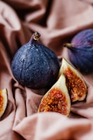 Two whole figs and a fig cut in half are placed on a soft, brown fabric. The figs have a rich purple color with a few droplets of water on their surface, suggesting freshness. The interior of the cut fig reveals a vibrant orange and red seeded flesh.