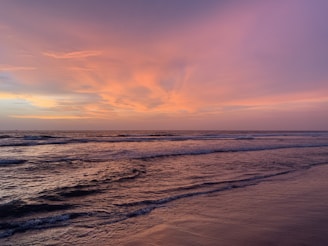A serene ocean view during sunset, with gentle waves lapping against the shore and the sky painted in warm hues of orange, pink, and purple. The horizon stretches far into the distance, creating a calm and tranquil atmosphere.